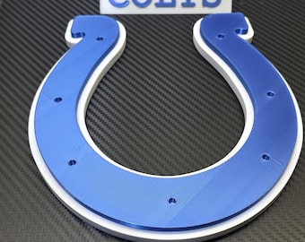 May include: A blue and white horseshoe-shaped logo with the word "COLTS" in white block letters. The logo is a 3D-printed design, likely for a sports team. The background is a dark, textured surface.