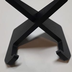 May include: A black, minimalist stand with an X-shaped top and two curved supports. The stand is made of a solid material and is designed to hold an item. The design is simple and modern.