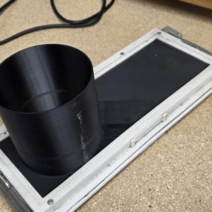 May include: A black cylindrical object sits atop a rectangular frame with a black interior. The frame has a silver border. A power cord with a plug is visible in the background. The items are on a light brown surface.