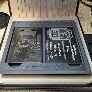 May include: A laser engraving machine with a business card/bottle opener design. The card size is 56 x 57 mm. The design includes the text "THEIRONREAPER" and "Black Swamp 3D". The machine has a fan and measurement markings.