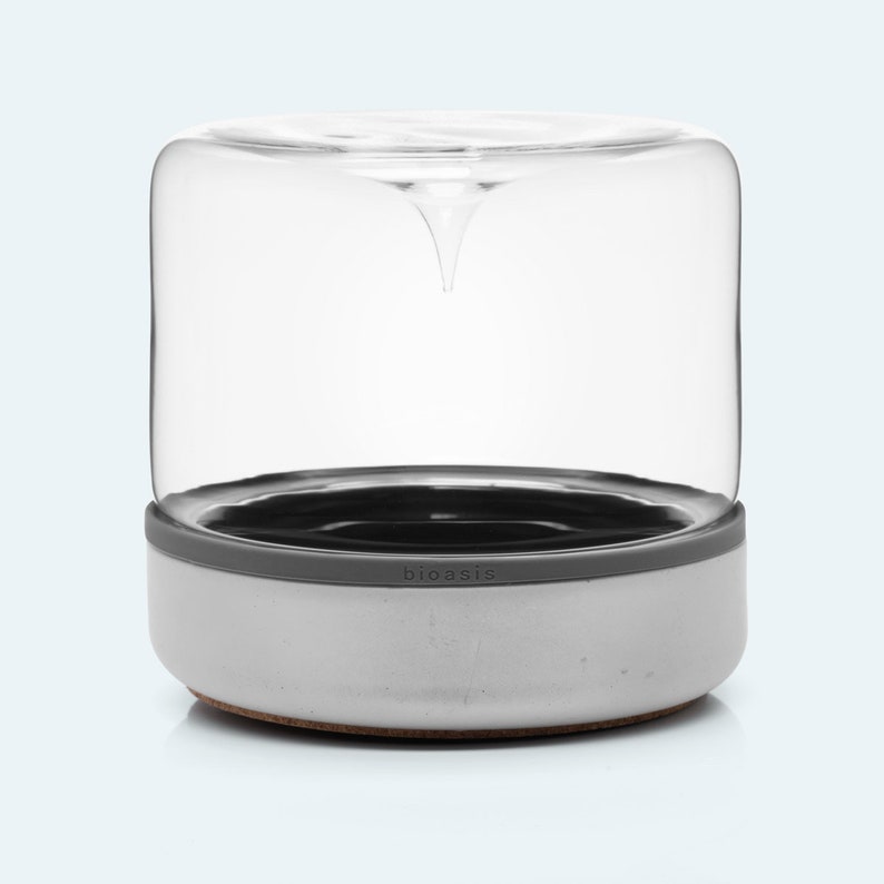 May include: A white ceramic planter with a gray silicone ring and a clear glass dome. The planter has the word "bioasis" printed on the side.
