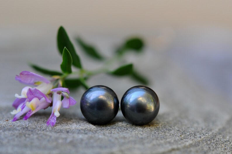 Puede incluir: Dos pendientes de perlas gris oscuro, sobre un fondo de una flor morada y hojas verdes.