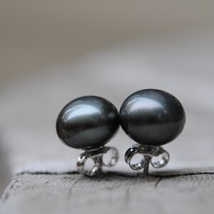 Puede incluir: Un par de pendientes de perlas negras con postes de plata. Las perlas son redondas y tienen un acabado liso y brillante.
