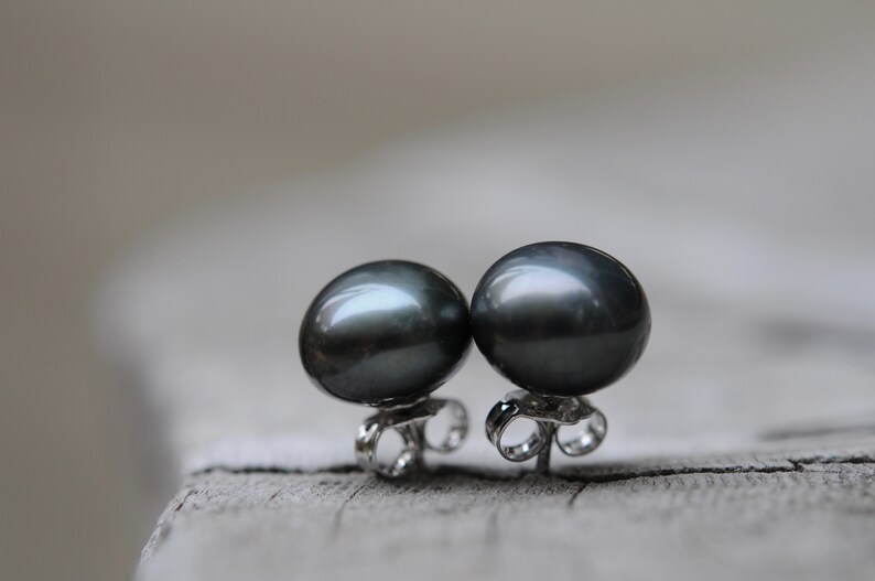 Puede incluir: Un par de pendientes de perlas negras con postes de plata. Las perlas son redondas y tienen un acabado liso y brillante.