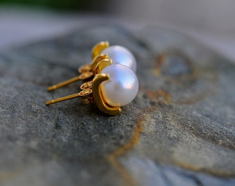 May include: A pair of gold pearl earrings with a simple design. The pearls are white and round.