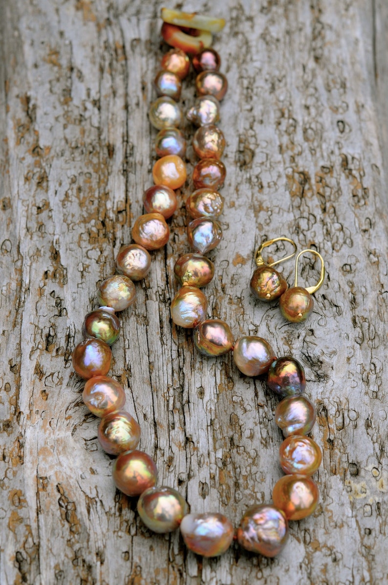 K&ouml;nnte beinhalten: Eine Kette aus irisierenden Perlen in den Farben Pfirsich, Rosa und Gold mit passenden Ohrringen. Die Perlen sind auf einer goldenen Kette aufgef&auml;delt und auf einer verwitterten Holzoberfl&auml;che pr&auml;sentiert.