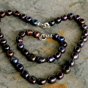 May include: A set of two necklaces made of dark iridescent pearls. The necklace is longer than the bracelet. Both pieces have silver clasps.