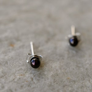 May include: Two small stud earrings with dark, possibly black, round gemstones. The earrings have a silver-colored post and backing. The studs are set against a light gray, textured surface.