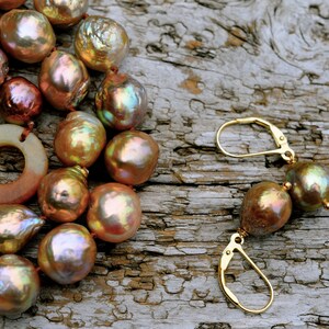 K&ouml;nnte beinhalten: Eine Halskette und Ohrringe aus irisierenden braunen Perlen. Die Halskette hat einen gro&szlig;en, flachen Anh&auml;nger aus braunem und wei&szlig;em Stein. Der Schmuck wird auf einer rustikalen Holzoberfl&auml;che pr&auml;sentiert.
