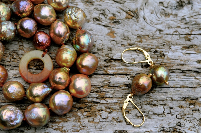 K&ouml;nnte beinhalten: Eine Halskette und Ohrringe aus irisierenden braunen Perlen. Die Halskette hat einen gro&szlig;en, flachen Anh&auml;nger aus braunem und wei&szlig;em Stein. Der Schmuck wird auf einer rustikalen Holzoberfl&auml;che pr&auml;sentiert.