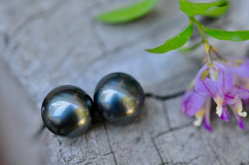 Puede incluir: Dos pendientes de perlas gris oscuro sobre una superficie texturizada gris claro con una flor morada en el fondo.