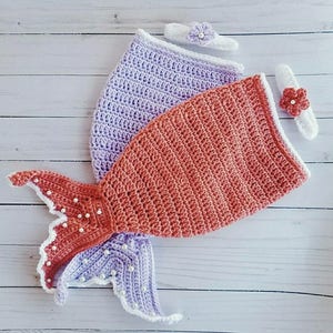 May include: A crocheted mermaid tail costume for a baby, featuring a purple and orange tail with white trim and pearl accents. The costume comes with two matching headbands with flowers.