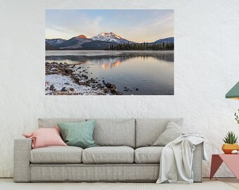 May include: A panoramic photograph of a snow-capped mountain reflected in a still lake. The mountain is surrounded by trees and the sky is a soft blue with hints of pink and orange.