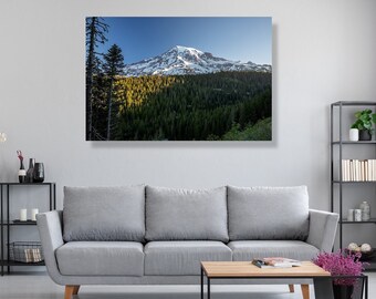 May include: A framed canvas print of a mountain landscape. The image features a snow-capped mountain peak, lush green forest, and a clear blue sky. The print is displayed above a gray sofa in a modern living room setting.
