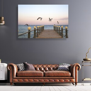 May include: A wooden pier extends out over a calm blue ocean with seagulls flying overhead. The pier has a railing with light posts and the sky is a soft blue with a hint of pink.