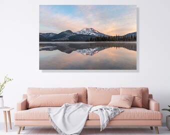 May include: A scenic photograph of a mountain reflected in a still lake at sunrise. The mountain is snow-capped and the sky is a soft pink and blue. The lake is calm and the water is reflecting the colors of the sky.