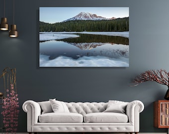 May include: A photograph of a mountain reflected in a lake. The mountain is covered in snow and trees. The lake is partially frozen and has a blue-green color. The sky is a light blue with some clouds.