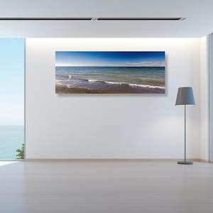 May include: A panoramic photograph of a calm, blue ocean with waves gently lapping at the sandy shore. The sky is a clear blue with a few wispy clouds. The image is framed and hanging on a white wall in a modern living room.