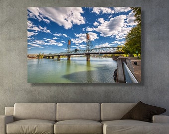 May include: A photograph of a bridge over a river with a blue sky and white clouds in the background. The bridge is a steel truss bridge with a green and blue color scheme. The river is calm and reflects the sky and clouds.