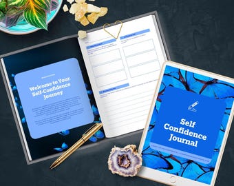 May include: An open journal and a tablet with the text "Self Confidence Journal" on a blue background with butterfly graphics. A pen, a decorative stone, and a plant in a decorative bowl are also visible.