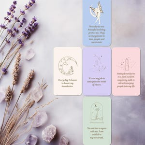 May include: A set of oracle cards with motivational quotes and drawings. The cards are displayed on a light grey surface with lavender and crystals. The cards have text including "Boundaries are beautiful and they protect me."