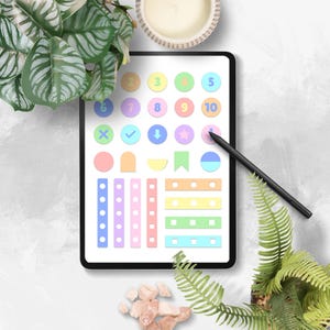 May include: A digital tablet displays colorful planner stickers, including numbered circles, checkmarks, arrows, and geometric shapes. A black stylus rests on the screen. The scene is decorated with greenery, a candle, and small pink stones.
