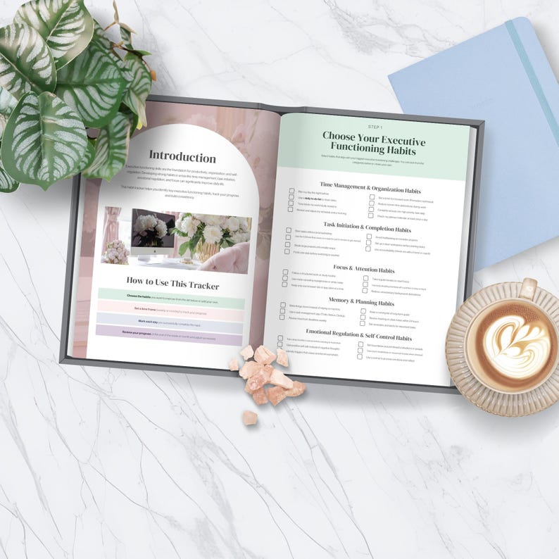 May include: An open planner with the title "Introduction" and "Choose Your Executive Functioning Habits" is displayed. A cup of coffee with latte art and a light blue notebook are on the marble surface. The planner has check boxes.