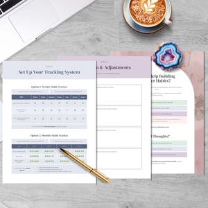 May include: A top-down view of a workspace with a laptop, coffee cup, and habit tracker printables. The printables include a weekly and monthly habit tracker, and a page titled "Help Building Better Habits?" A gold pen rests on the pages.
