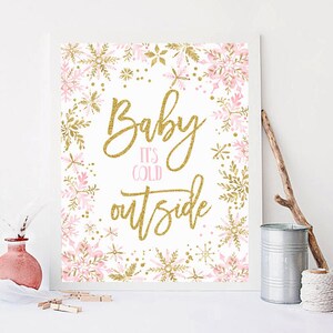 Puede incluir: Un cartel imprimible rosa y dorado con el texto "Baby It's Cold Outside". El cartel está decorado con copos de nieve y un fondo de purpurina dorada.