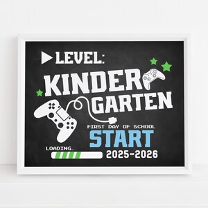 May include: A framed chalkboard-style sign with white text and graphics. The sign reads "LEVEL: KINDERGARTEN" with a video game controller graphic and the words "FIRST DAY OF SCHOOL START 2025-2026". A loading bar graphic is also present.
