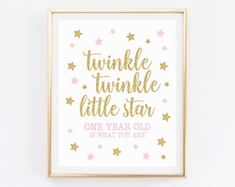 May include: Gold glitter framed print with the text "twinkle twinkle little star one year old is what you are" with pink and gold stars scattered around the text.