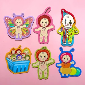 May include: A collection of six colorful stickers featuring baby-like figures in various costumes. Designs include a butterfly, cherry, clown, a basket of babies, an apple, and a caterpillar. The stickers are on a pink background.