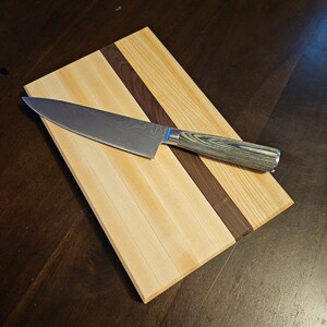 May include: A wooden chopping board with a light brown wood and a dark brown wood inlay. A silver and black chef's knife is placed on the board.