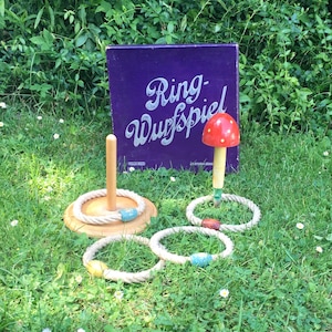 Op de afbeelding: Een vintage ringwerpspelset op gras. De set bevat een houten basis met een centrale pin, touwringen en een paddenstoelvormig doel. De doos op de achtergrond leest "Ring-Wurfspiel" in witte letters op een paarse achtergrond.