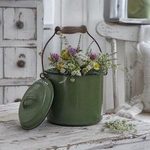 Pode incluir: Um balde de esmalte verde-oliva com uma alça de madeira e uma tampa, cheio de um buquê de flores silvestres. O balde é verde-escuro e a tampa está ao lado. As flores incluem flores amarelas, roxas e brancas.