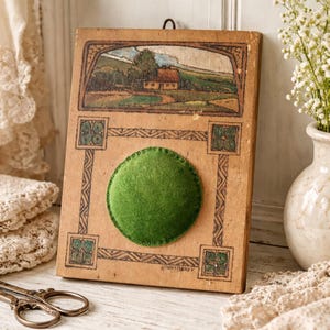 May include: A vintage wooden pin cushion with a green velvet centre. The wooden frame features a painted landscape scene and decorative corner designs. A metal loop is attached for hanging.