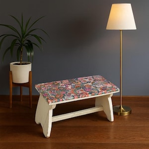 Puede incluir: Un pequeño banco de madera blanco con un asiento estampado con motivos de cachemira de colores. El banco está sobre un suelo de madera oscura, con una lámpara de pie alta y una planta en maceta en el fondo. El banco mide unos 76 cm de largo.