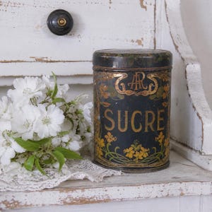 May include: A cylindrical black metal canister with a removable lid, featuring the word SUCRE in gold lettering, an ornate gold monogram, and yellow floral illustrations. The container has a weathered finish and sits next to white flowers.