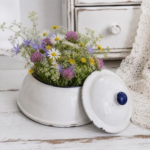 May include: A white enamelware bowl brimming with colourful wildflowers, including daisies, purple clover, and yellow flowers. The bowl has a matching lid with a blue knob. It rests on a distressed white wooden surface.