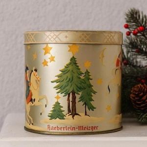 Peut inclure: Une boîte ronde vintage avec un fond argenté et un bord doré. La boîte présente des illustrations de sapins de Noël, d'étoiles et de personnages à cheval. Le texte "haeberlein-metzger" est imprimé en bas.