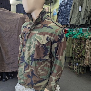 May include: A camouflage jacket with a button-up front and multiple pockets. The jacket is displayed on a mannequin wearing a white skirt.