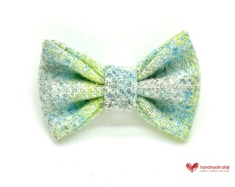 May include: A small, colorful bow tie with a tweed-like texture. The bow tie features a mix of light blue, green, and gray threads, creating a textured appearance. The bow tie is likely for a dog, as indicated by the logo in the bottom right corner.