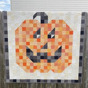 May include: A patchwork quilt featuring a large orange and black jack-o'-lantern design. The quilt is made of various fabrics in shades of orange, black, and white. The jack-o'-lantern has a wide, toothy grin and a pair of large, black eyes.