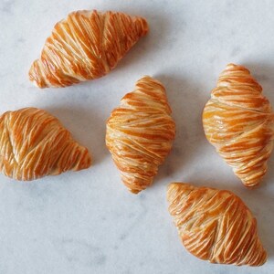 Puede incluir: Cinco croissants de color marrón dorado dispuestos sobre una superficie de mármol blanco.