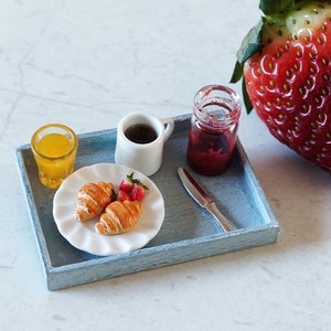 Könnte beinhalten: Miniatur-Frühstücksszene auf einem blauen Tablett. Enthält ein kleines Glas Orangensaft, eine weiße Kaffeetasse, ein Glas Marmelade, einen Teller mit Croissants und Erdbeeren sowie eine große Erdbeere. Das Tablett hat auch ein kleines Messer.