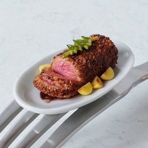 Op de afbeelding: Een miniatuur bord met een klein, gesneden en gepaneerd stuk vlees, een paar kleine, ronde, gele aardappelen en een groene garnering. Het bord ligt op een zilveren vork.