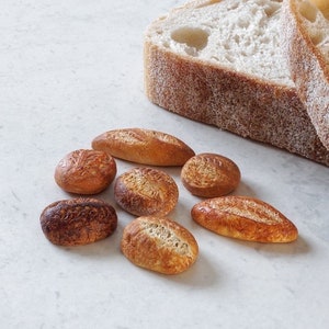 Puede incluir: Una variedad de pequeños panes y panecillos, incluyendo baguettes y panecillos redondos, están dispuestos sobre una superficie blanca. Dos rebanadas de pan crujiente están en el fondo. El pan tiene una corteza dorada y un interior claro. La imagen incluye el logo "Sois Creations".