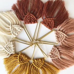 May include: A set of six pairs of handmade earrings featuring a variety of colors and textures. Each earring is made with a triangular gold-tone metal base and a tassel of yarn in a different color. The colors include cream, brown, peach, and gold.