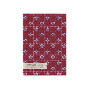 May include: A rectangular pack of scrapbooking papers with a deep red background featuring a grid of dashed light blue lines and small light blue flower icons. A beige label at the bottom reads PATCHWORK GARDEN SCRAPBOOKING PAPERS in black text.