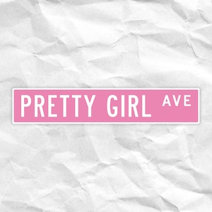 Puede incluir: Un letrero de calle rosa con las palabras "PRETTY GIRL AVE" en letras blancas. El letrero es rectangular y está sobre un fondo blanco texturizado.